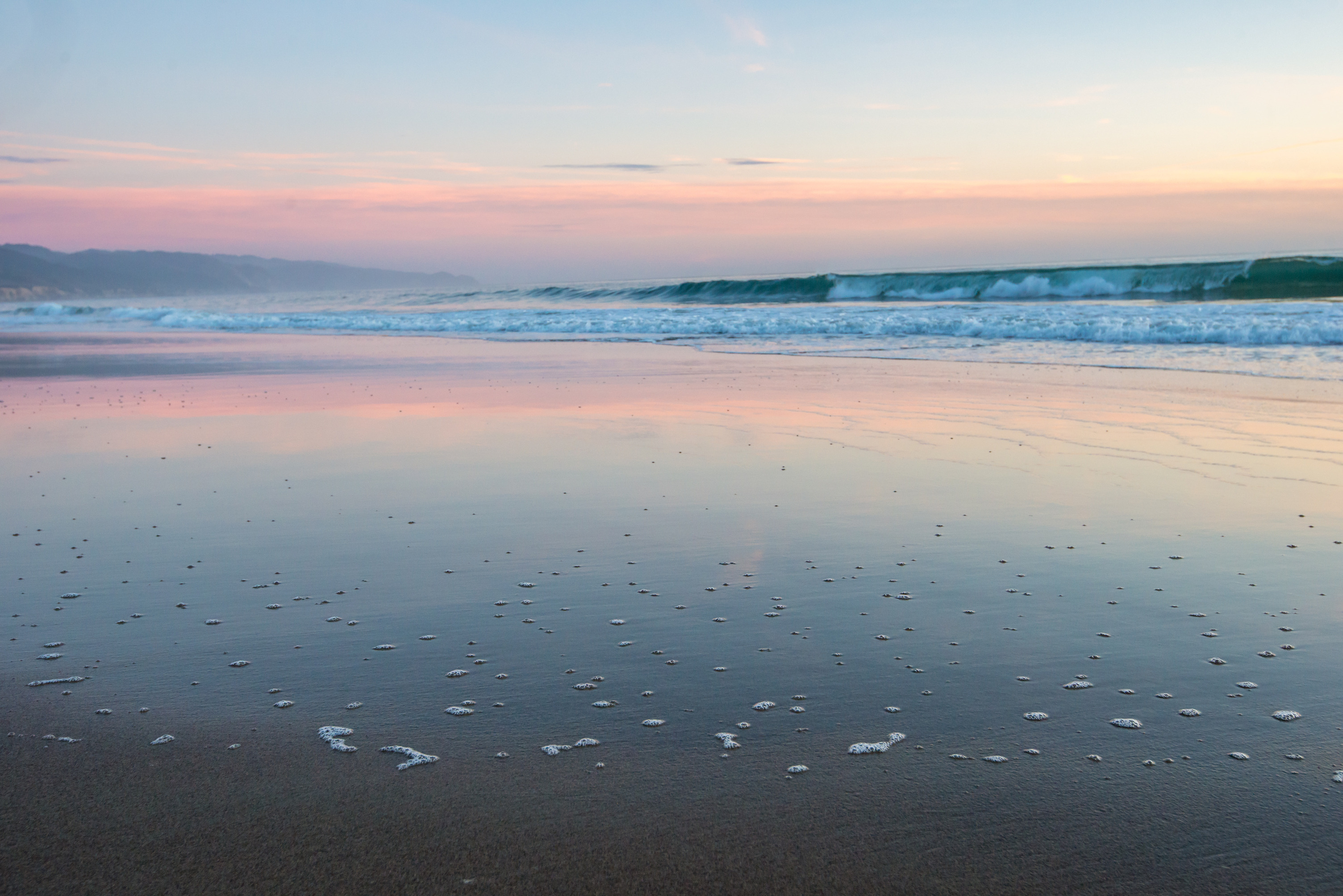 Pastel sunset reflected in wet sand dotted with clusters of bubbles from the previous wave as another wave crashes offshore.