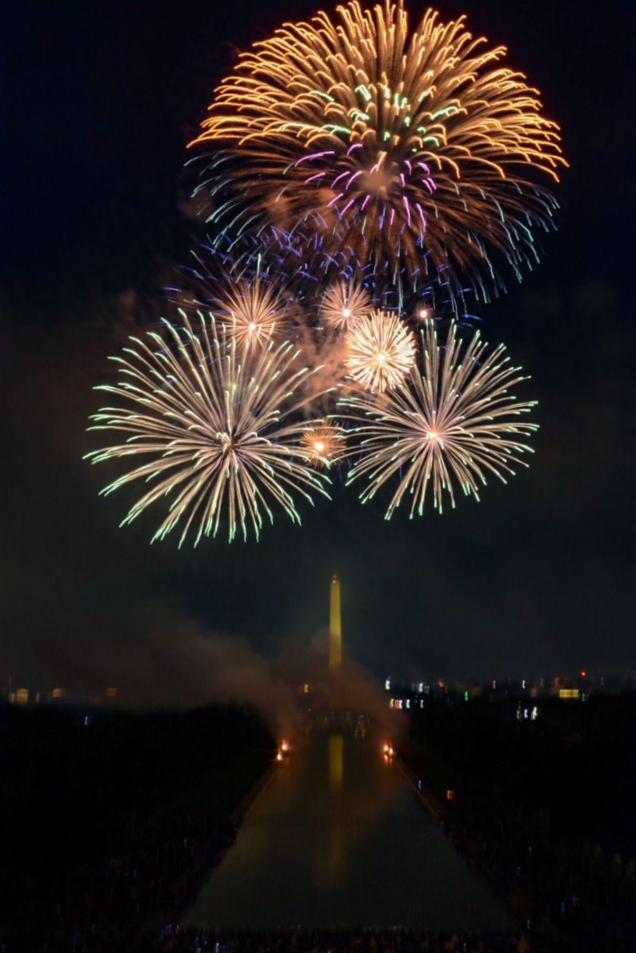 Colorful fireworks exploding above a reflecting pool at night, leading towards the Washington Monument.