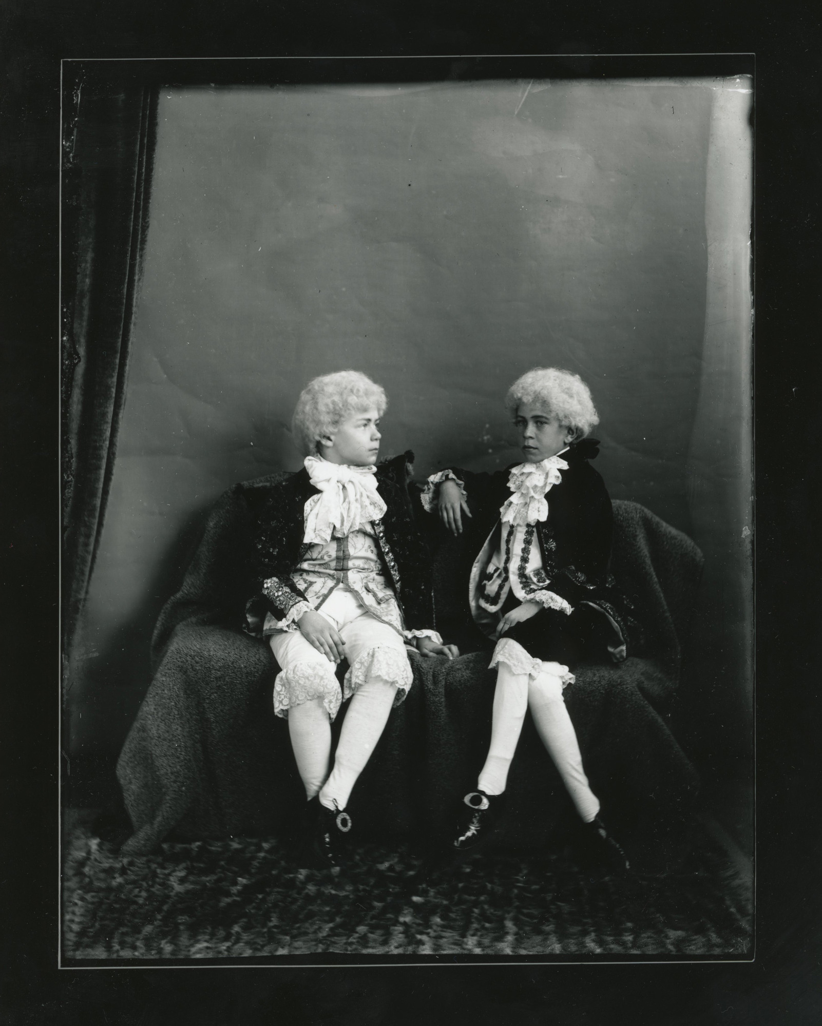 Two young white boys sit leaning towards each other. They wear full colonial costume.