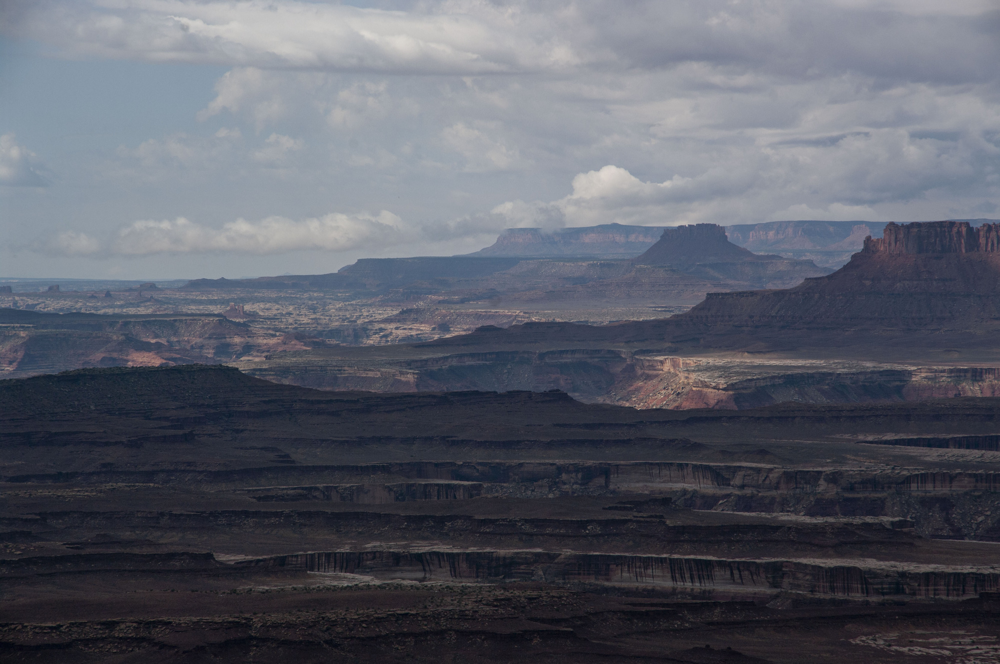 Shadowed canyons make up the foreground. In the background orange, sandstone sheer cliffs stand on a cloudy day. 