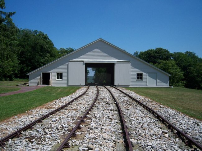 A white structure with a gable roof. The door of the Engine House are open and there is railroad track leading up to it.