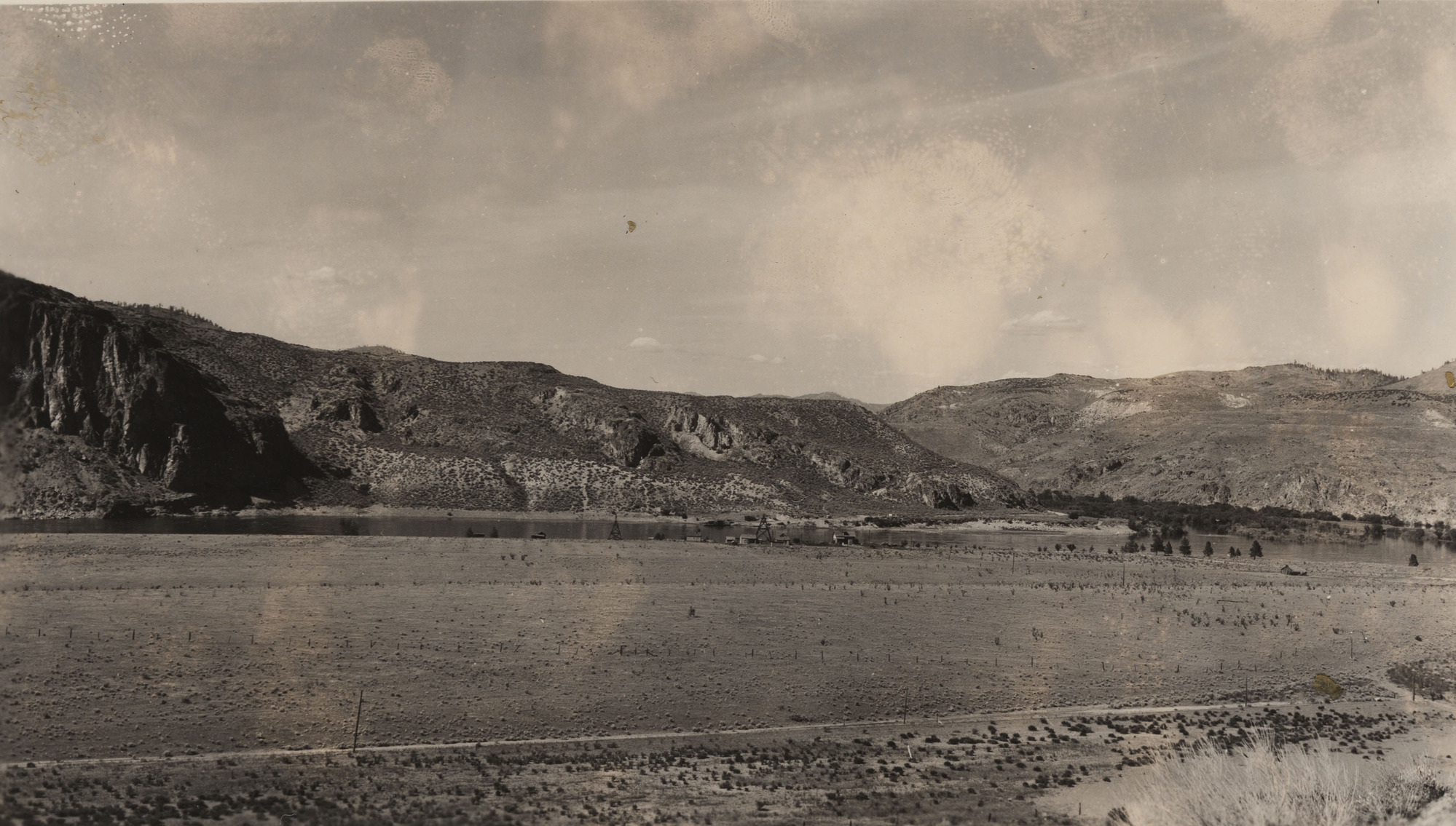 Slightly faded black and white photograph of a wide, bare floodplain and a river, with steep, rocky hills on the far side of the river