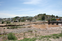 
A muddy river flowing under a bridge with a view of a hilly neighborhood and clear skies in the background.