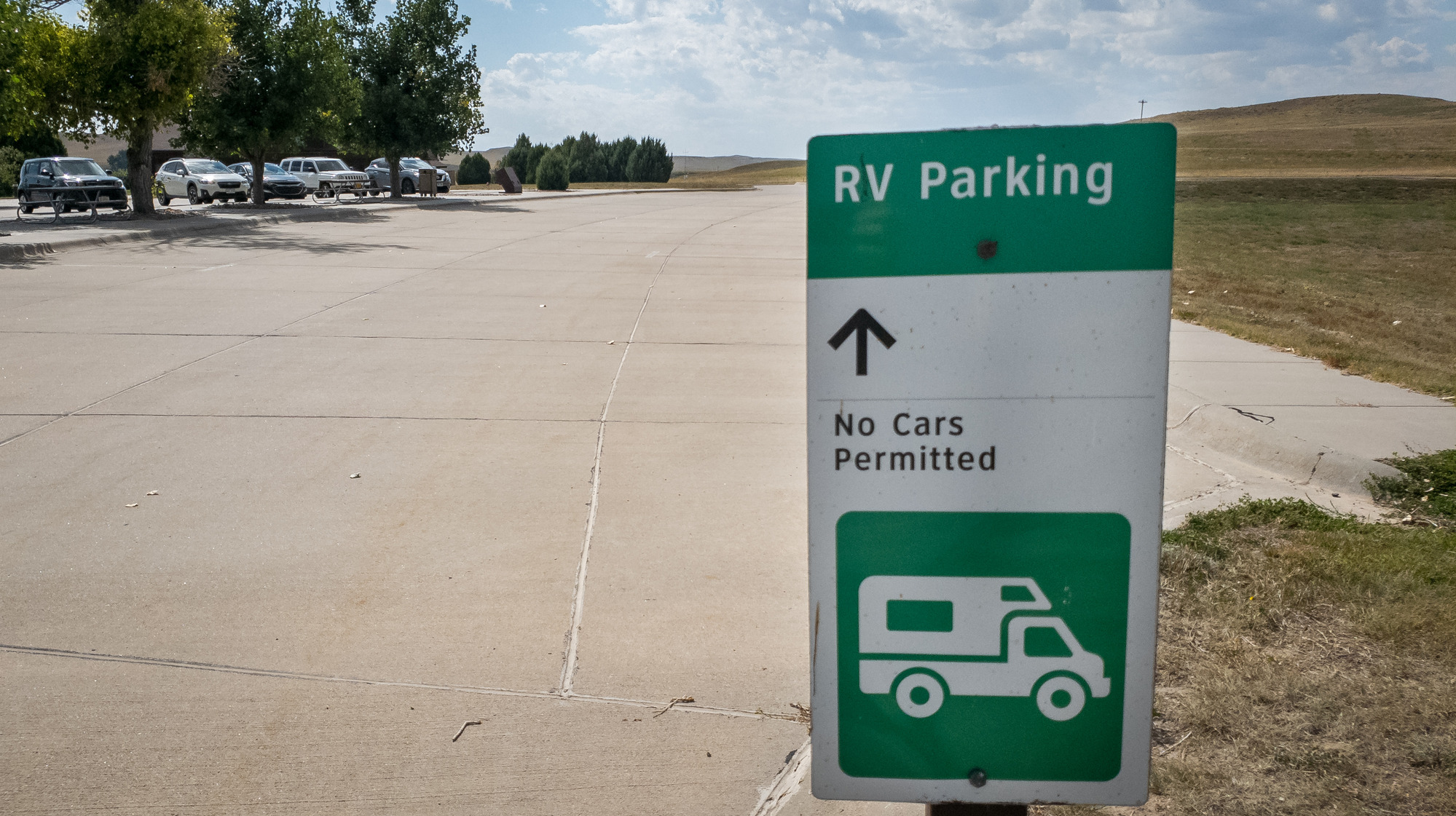 Green and white sign reads RV Parking No Cars Permitted in front or a large empty concrete parking lot and green trees.