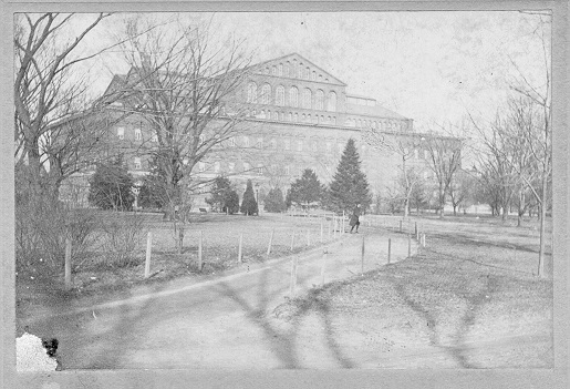 Photo is faded with some damage on the left bottom corner.  A pathway leads up to the building.  A person is crossing the path.  A variety of trees are in the photo around and in front of the building.