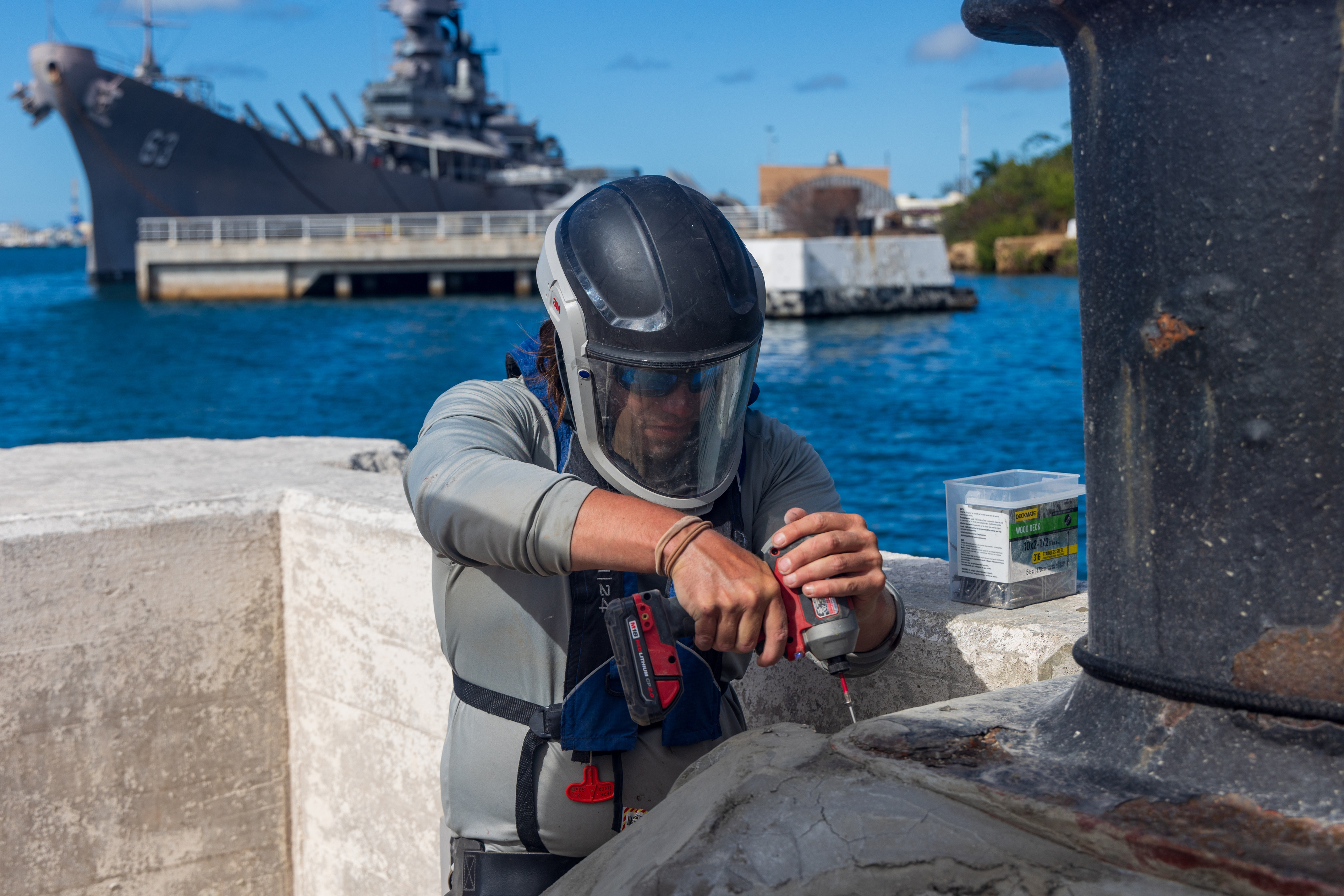 Man in protective headgear uses compact drill to drill into cement of mooring quay, harbor and floating battleship in background.