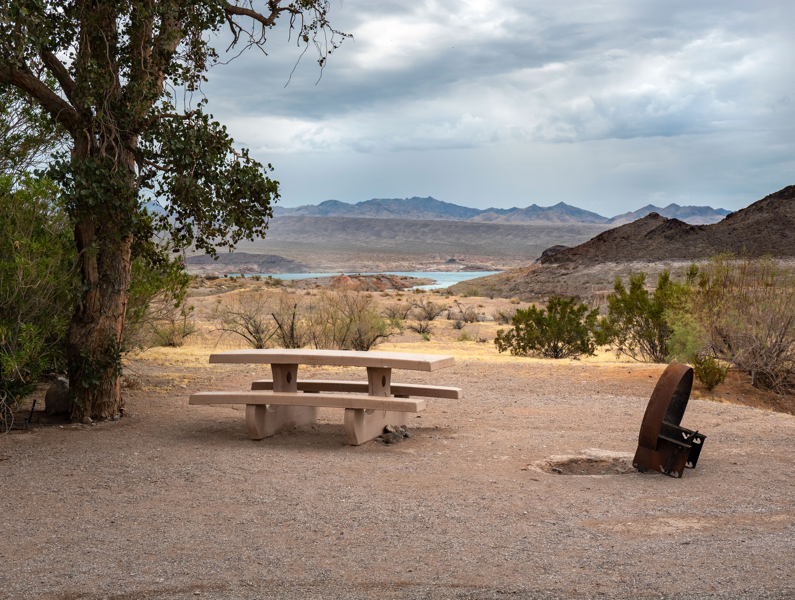 Concrete picnic bench, grill, large tree on left, mountains and body of water seen in distance