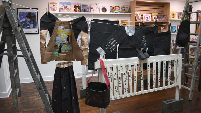 Clothing made out of brown paper painted black hangs from a clothesline including a jacket, pants, hat, and shirt. A tall skirt made out of brown paper painted black with red spots stands next to a picket fence with names painted in black on each picket atop the fence is a pair of brown paper shoes painted black and hanging from the fence is a black basket.