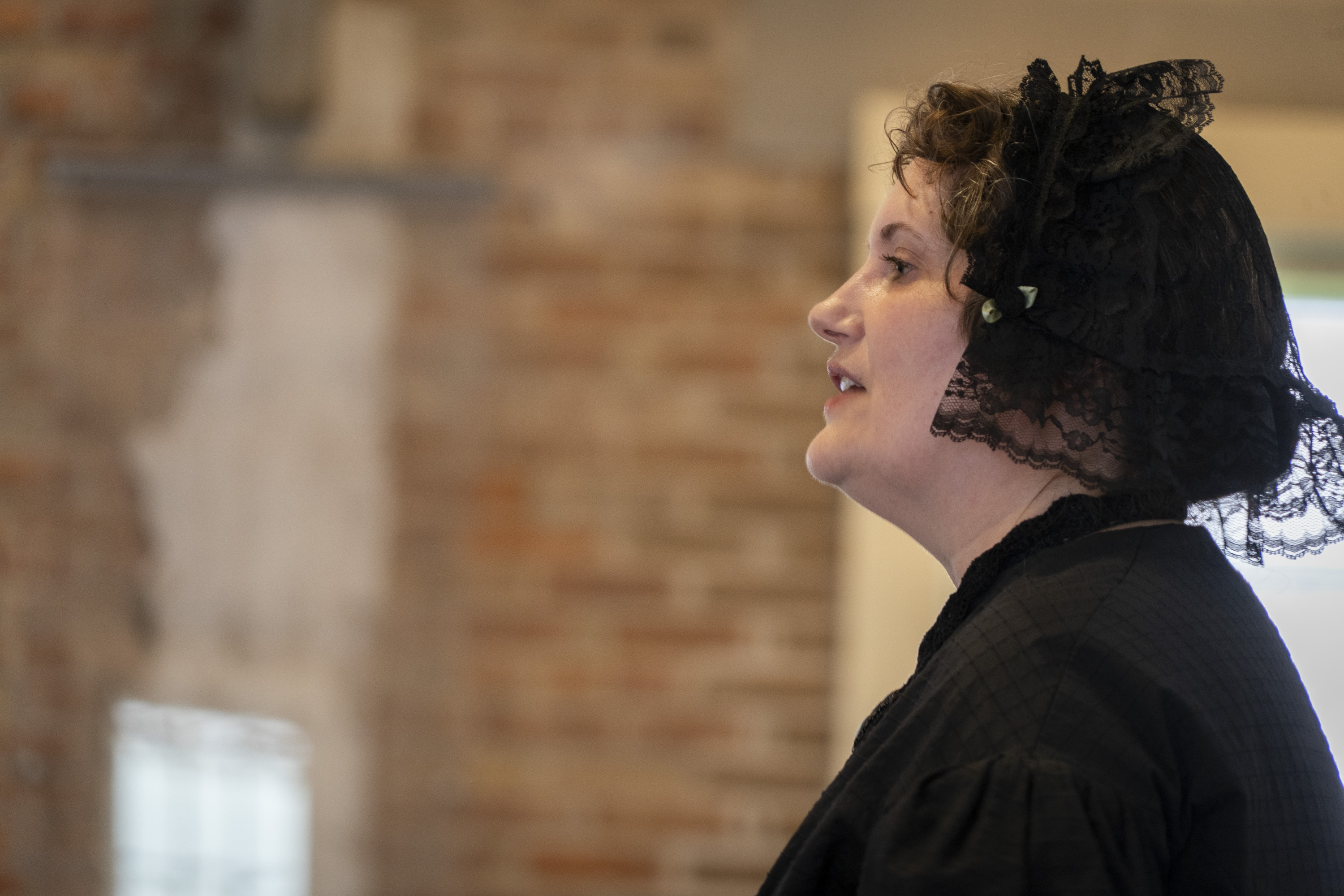 A woman in 19th century clothing speaks inside a brick room.