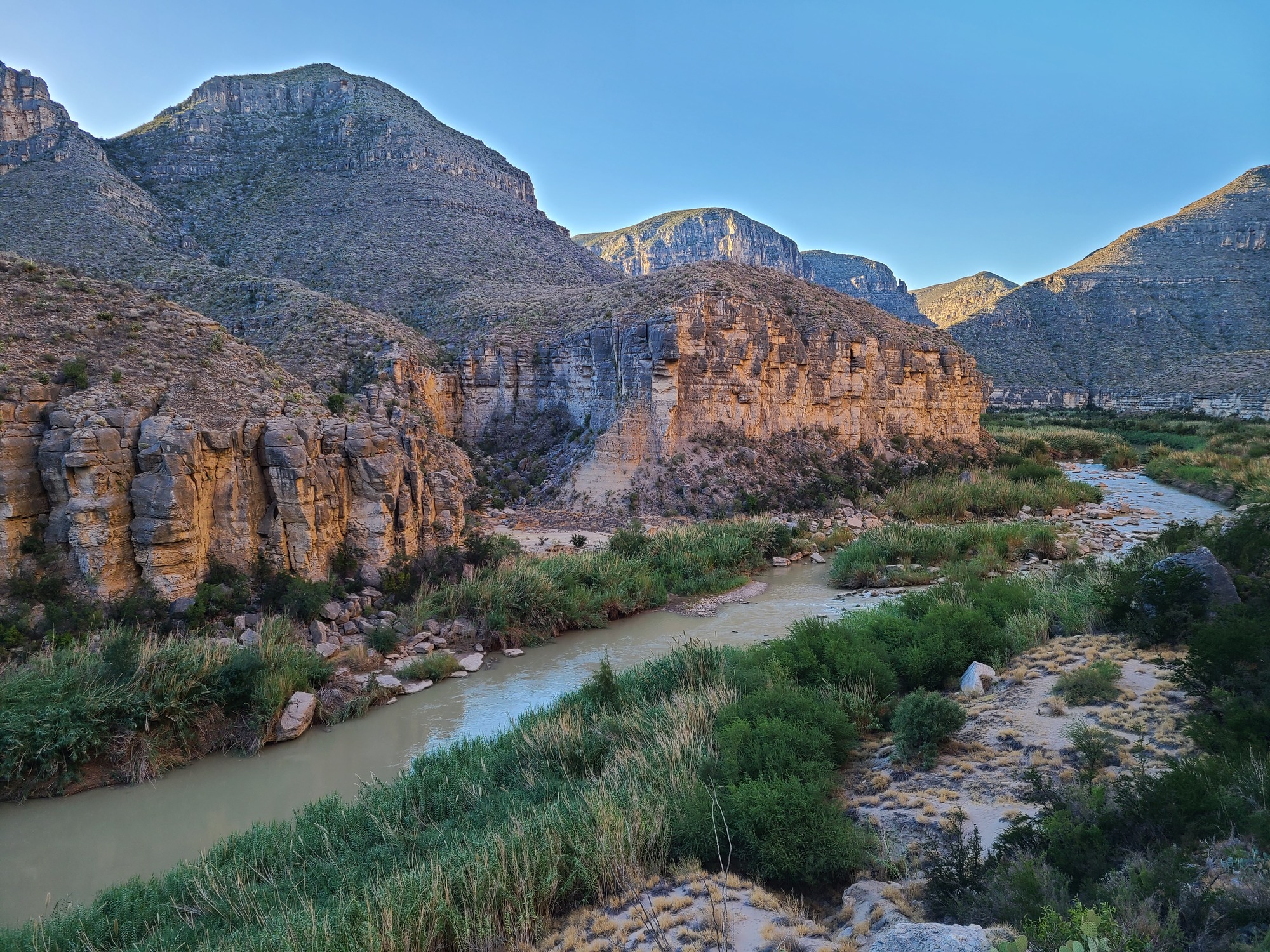 A view of the Rio Grande from a cliff above the river. From top to bottom, the image shows blue cloudless sky, steep beige cliffs dotted with green desert vegetation, a silty-green river interrupted by a rock garden rapid and in island, and bright green giant cane growing on both sides of the channel.