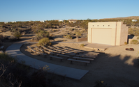 A small outdoor amphitheater with six rows of benches and a small stage area with a small building behind it. There is a fire pit to the right side. 