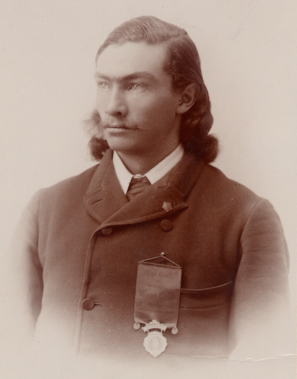 A sepia-toned photograph of a young man with long hair and a mustache. He is wearing a double-breasted coat with a metal medal or badge attached to a ribbon; the ribbon has text that reads in part "Chief Guide Wonderful Wind Cave".