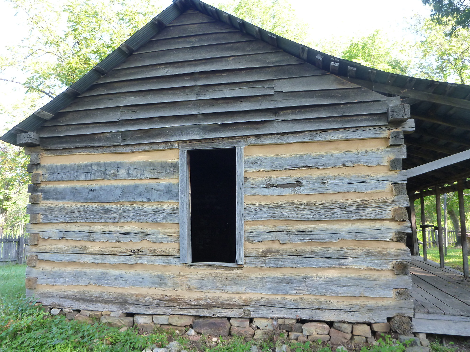 stacked rocks and green grass at bottom, grey logs with tan chinking and window opening at center, planks forming peaked roof at top
