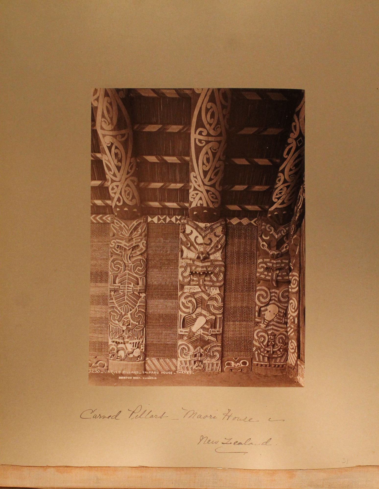 Interior of Maori house showing three elaborately carved wooden pillars. Each pillar has a representation of a fierce-looking figure with protruding tongue.