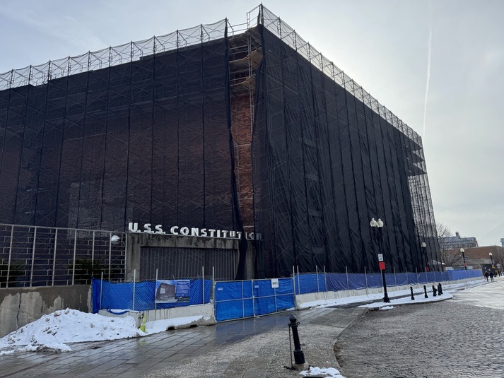 Scaffolding covered in black scrim surrounds Hoosac Stores, a red brick industrial building. A fence with blue scrim separates it from the sidewalk that runs alongside it. 