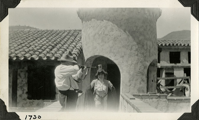 This is an historic black and white photograph from the Scotty's Castle Historic Photograph Collection, Death Valley National Park of Walter Edward Perry Scott pretending to shoot a can off of Eva Mudge's head, on Scotty's Castle Main House and Annex Bridge. Circa 1928. Photographed by Mat Roy Thompson.