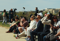 A group of people gather to watch and take photos of the partial eclipse on a large lawn