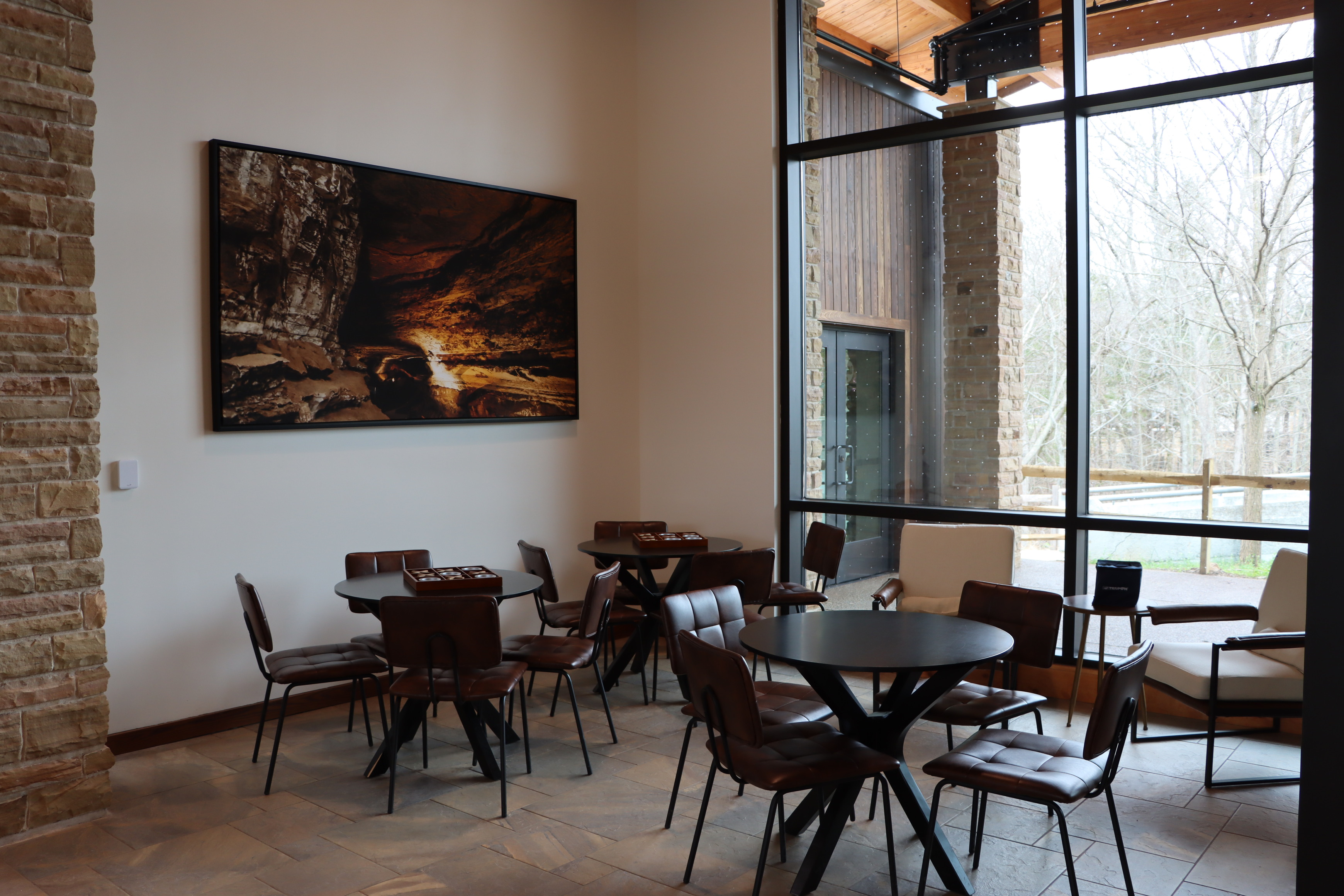 Several tables and chairs are arranged in a brightly lit corner with glass windows and a large photo of a cave passageway hanging on the wall.