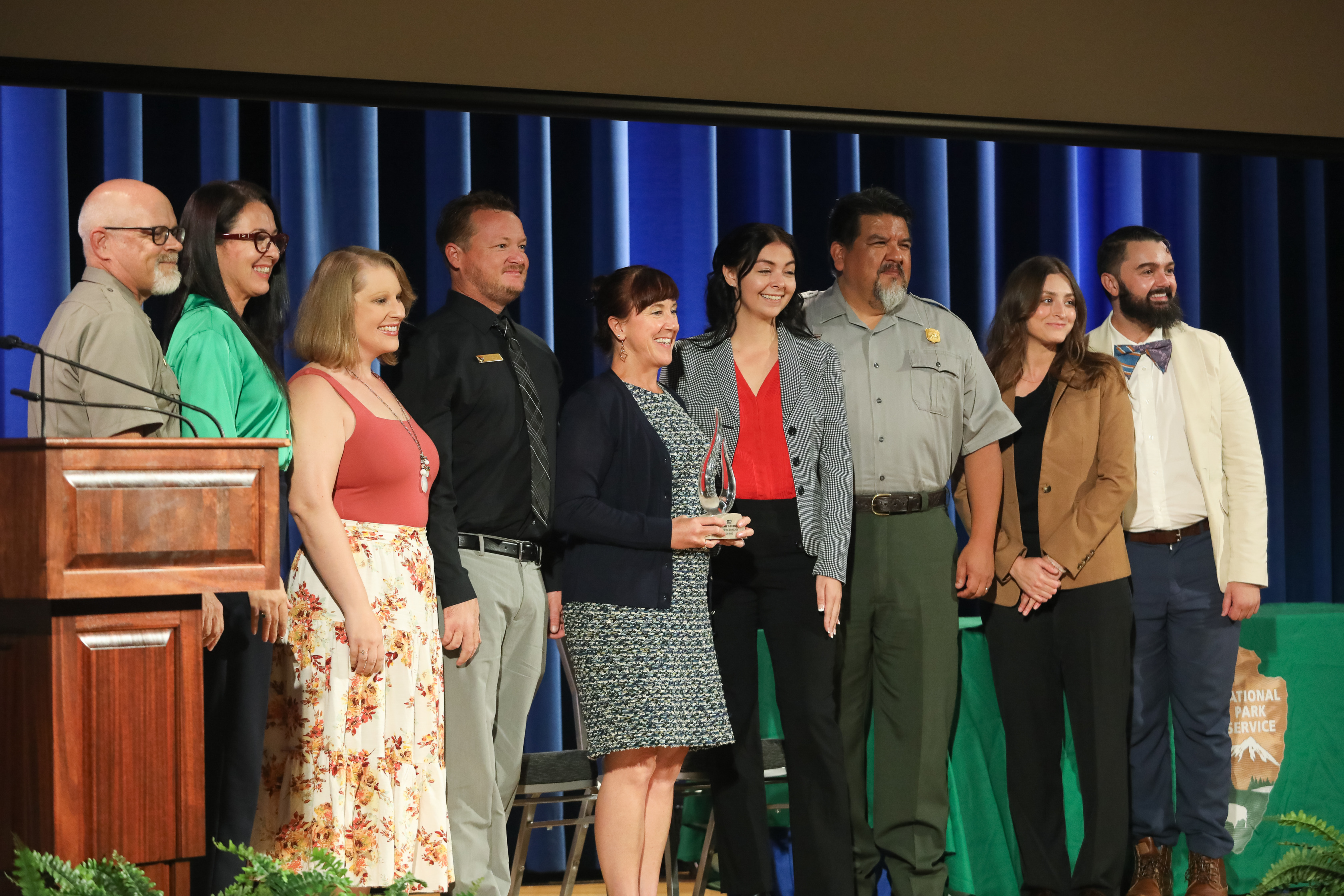 A group of 9 people look off camera while posing for a photo during an award ceremony.