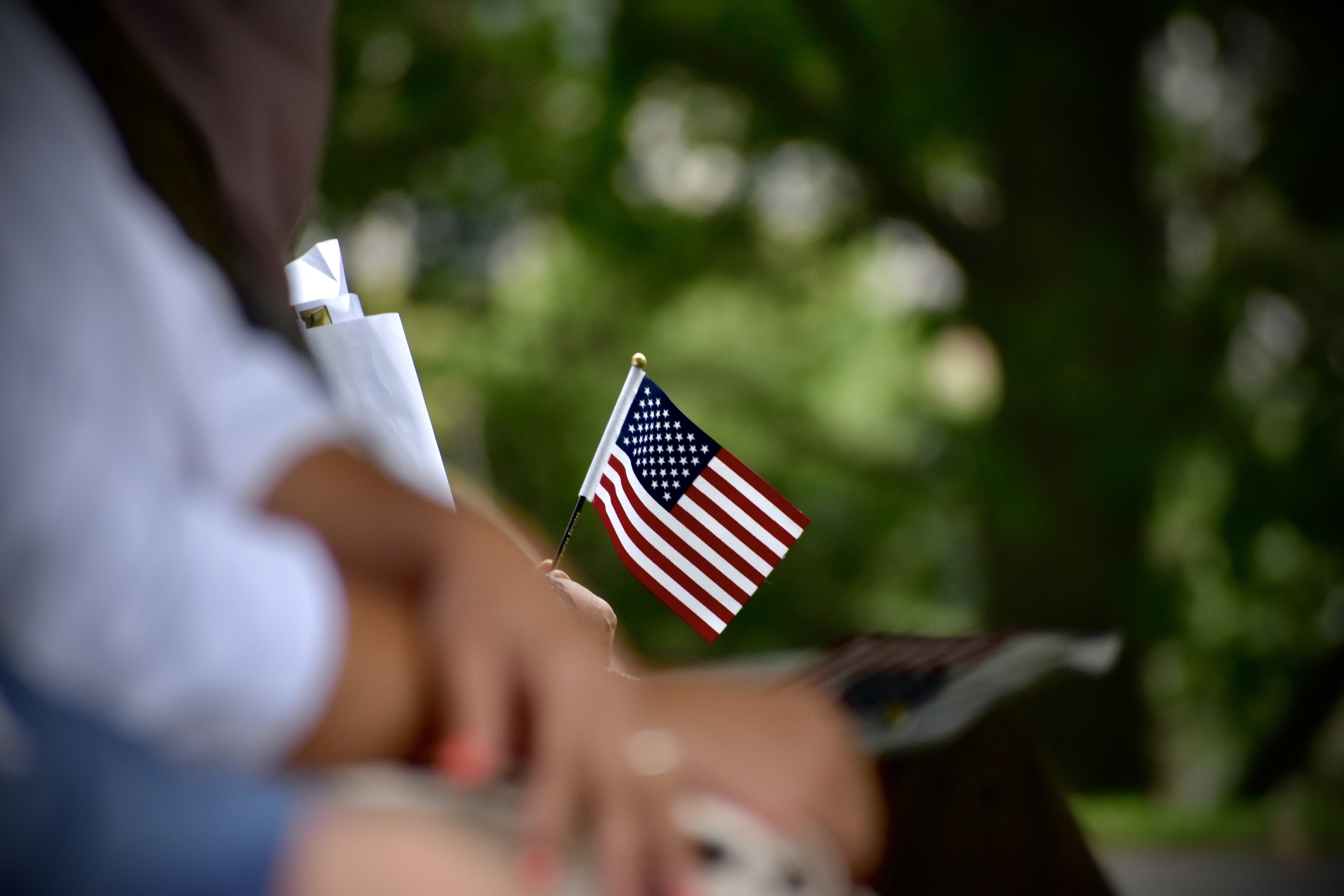 The American flags is being held by an individual who is seated. Their are hands folded in the foreground of the picture. 