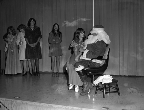 Christmas holiday party, 1976. Girl sits on Santa's lap on stage in Visitor Center auditorium, while others wait in line.
