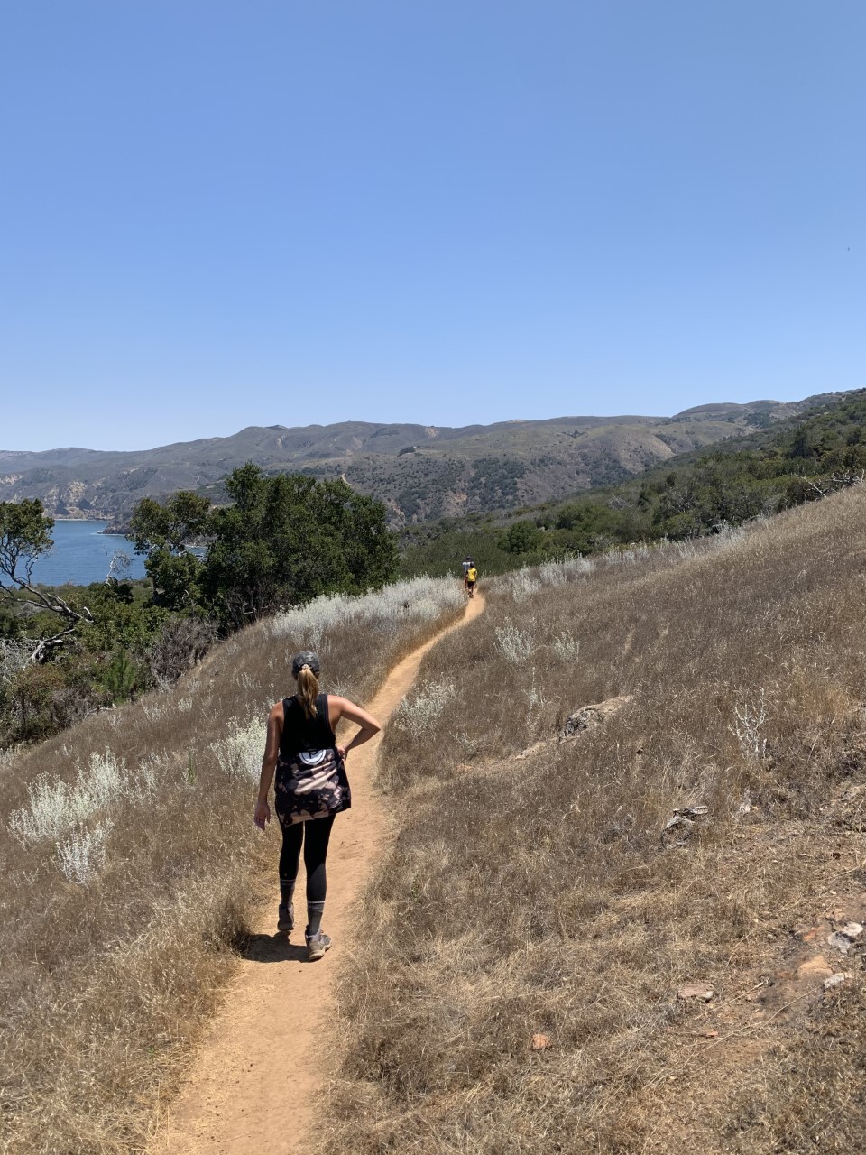 A woman in black leggings, a black sleeveless top and a gray baseball cap with a blonde pony tail sticking out looks down a narrow, dirt trail cut into a dry, grassy landscape where a child in a yellow shirt leads in the distance. Tall, bushy trees and a green mountain range pop up in the background with a small peak of blue ocean water.