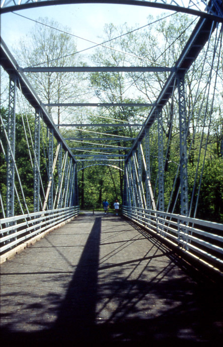  Two people walk down a metal truss bridge painted pale grey.