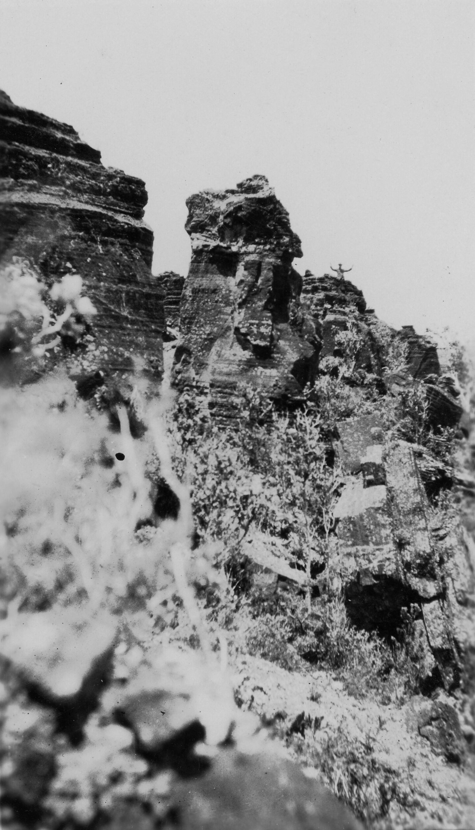 Black and white photograph showing a view from within a crater. The terrain is steep and jagged with many rocks. Vegetation can be seen along the rocks, but appear very dry. Towards the top half of the image, tall, vertical boulder formations extend upwards. Off-center, towards the right side, a person can be seen far in the distance on top of the boulders, close to the rim of the crater. The unidentified person poses for the camera with their arms in the air.