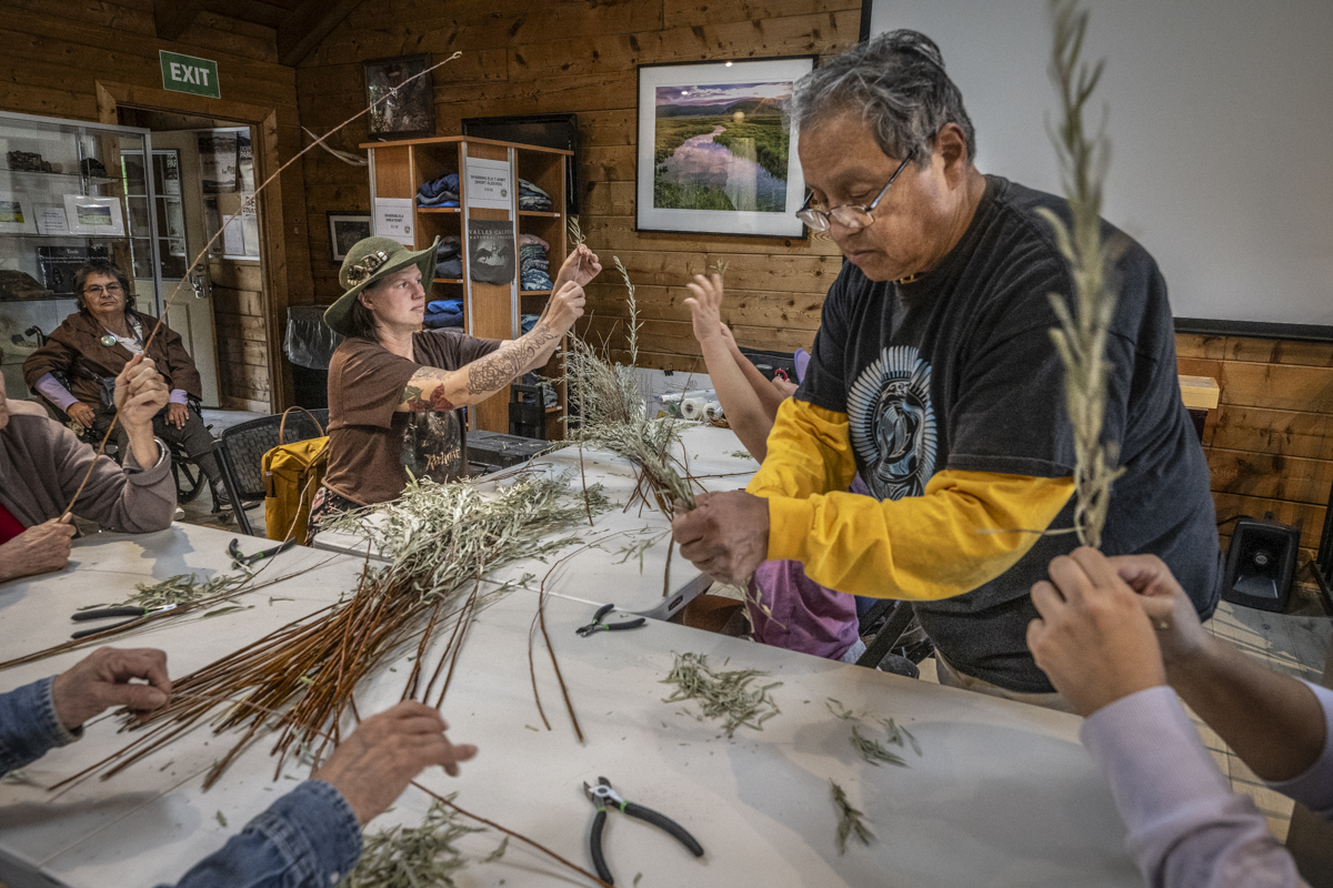 A man stands and removes leaves from a willow cutting while surrounded by seated participants doing the same.