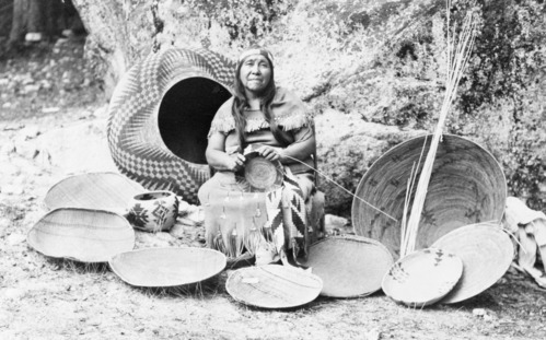 copied by Onas Ward. Lucy Telles posed with baskets, most of which she made. Large one on left is famous one in Yosemite Museum collection. Unfinished tray @ right also in Yosemite collection.