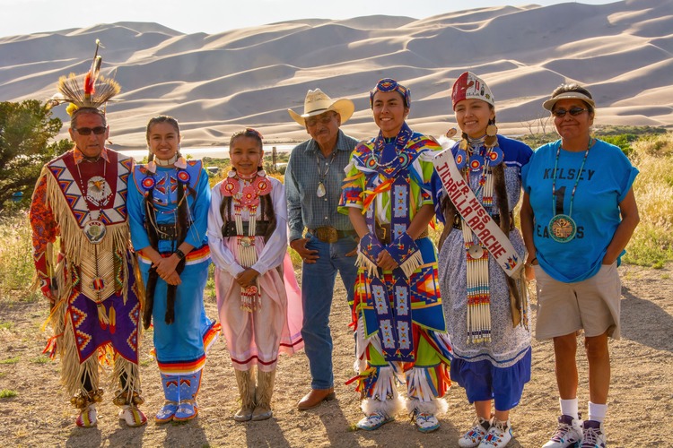 Wearing colorful regalia, the Jicarilla Apache dance team smiles in front of the dunes after performances at the park amphitheater. The dunes are illuminated by early evening sunlight with patterns of light and shadow.