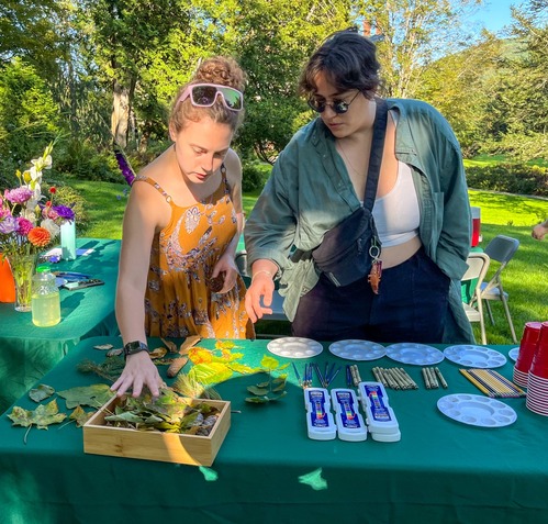two people look at table with leaves, paint pallets, paint, and paintbrushes in an outdoor setting