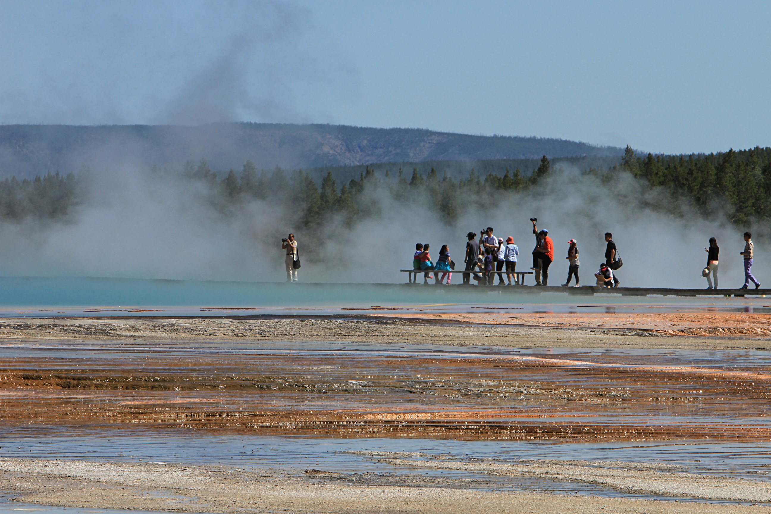 A group of people standing on a boardwalk behind a steaming hot spring that is reflecting the color blue into the steam.