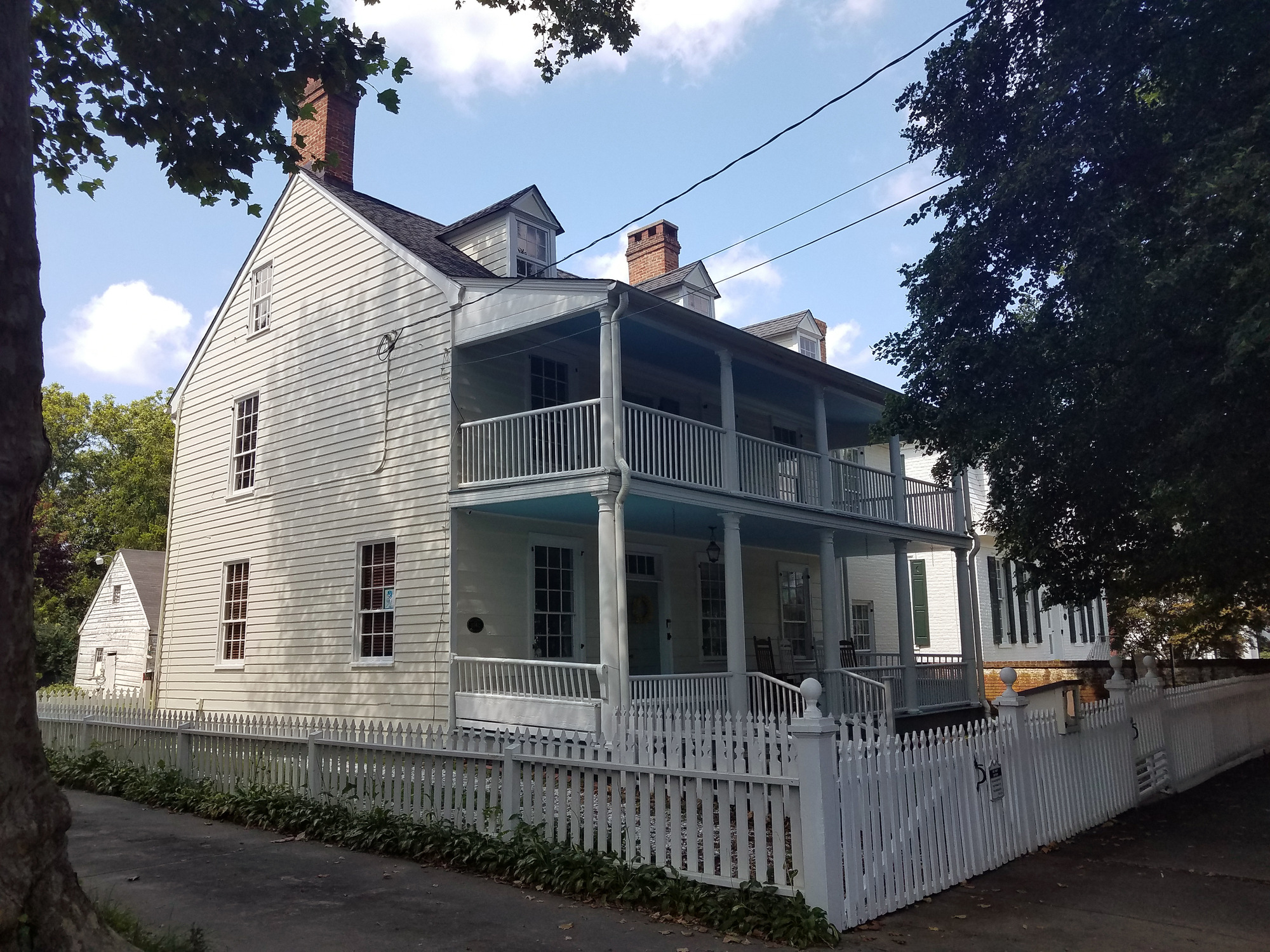 A three story, non-original, pale yellow house surrounded by a white picket fence. There is a porch and balcony on the front of the home, with three dormer windows and two chimneys on the roof. A white outbuilding peers out from behind the house.