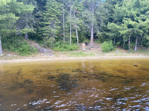 View of beach landing for houseboat site with the houseboat sign on edge of beach with fire ring farther up shore surrounded by trees. Large pine trees cover the background of image.