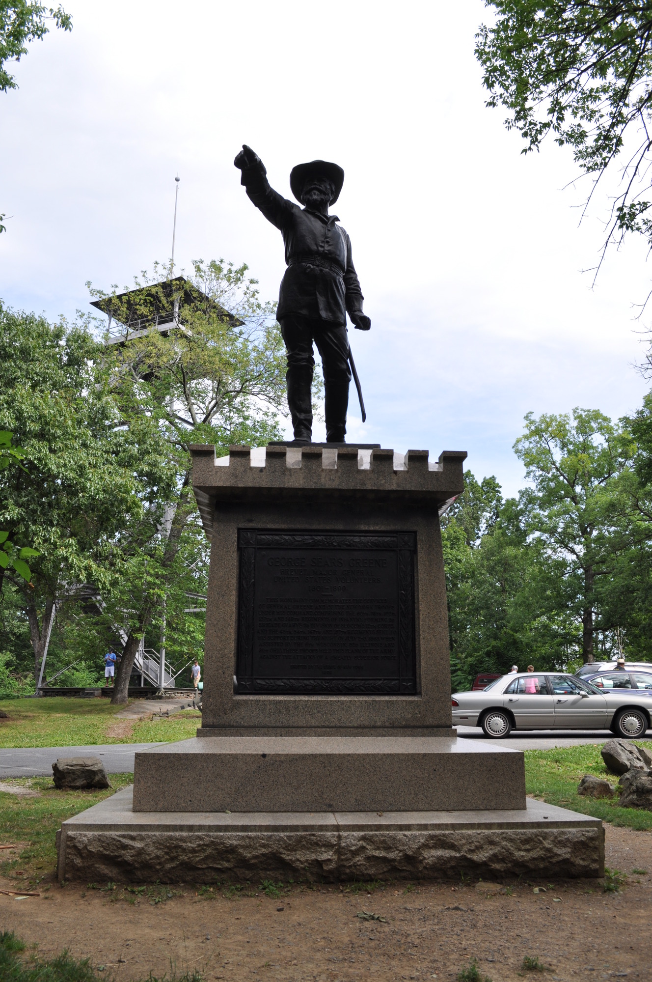 Statue of a man with hat and sword extending his right arm to point, atop a rectangular pedestal. Trees, a tall watchtower, and parking lot in the background.