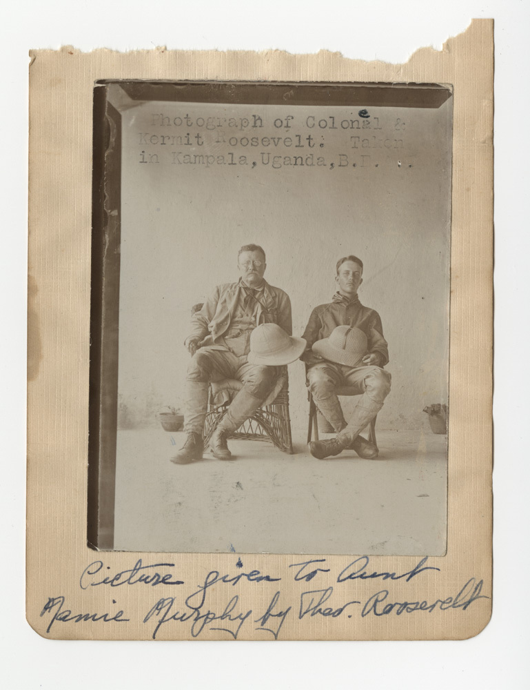 A father and his older son sit next to each other in chairs, posed seriously for the portrait. They hold pith helmets in their lap.
