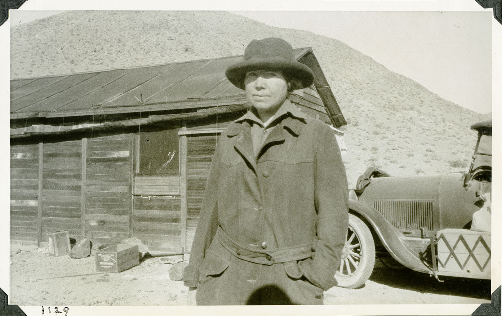 This is an historic black and white photograph from the Scotty's Castle Historic Photograph Collection, Death Valley National Park of a woman posing for the camera in front of a simple wood structure and passenger car. Woman in rolled-brimmed hat, shirt, and heavy coat. A few crates and rocks stacked next to building foundation. Desert hillside in background. Number in black ink in lower left corner.
