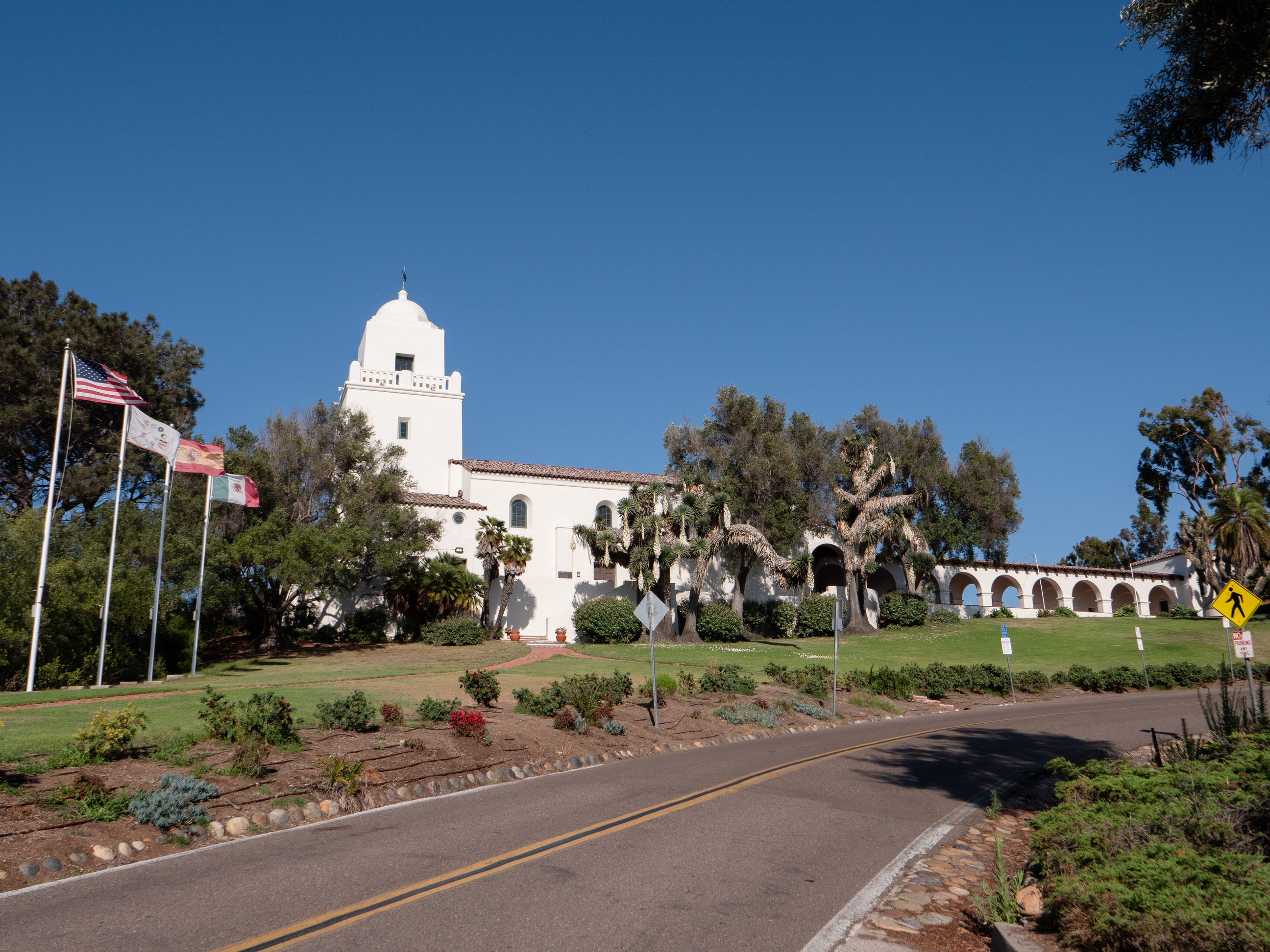 A long white building on a hillside has a covered walkway with many arches, a Spanish-style tiled roof, and a bell tower with four flags flying in front