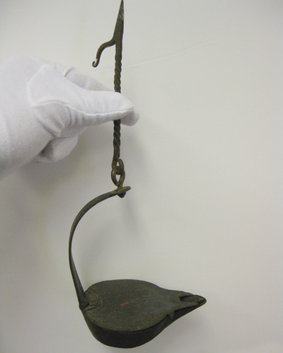 Curator's white gloved hand holds a small, iron hook, hanging from it is a curved iron band holding a small, covered iron bowl.