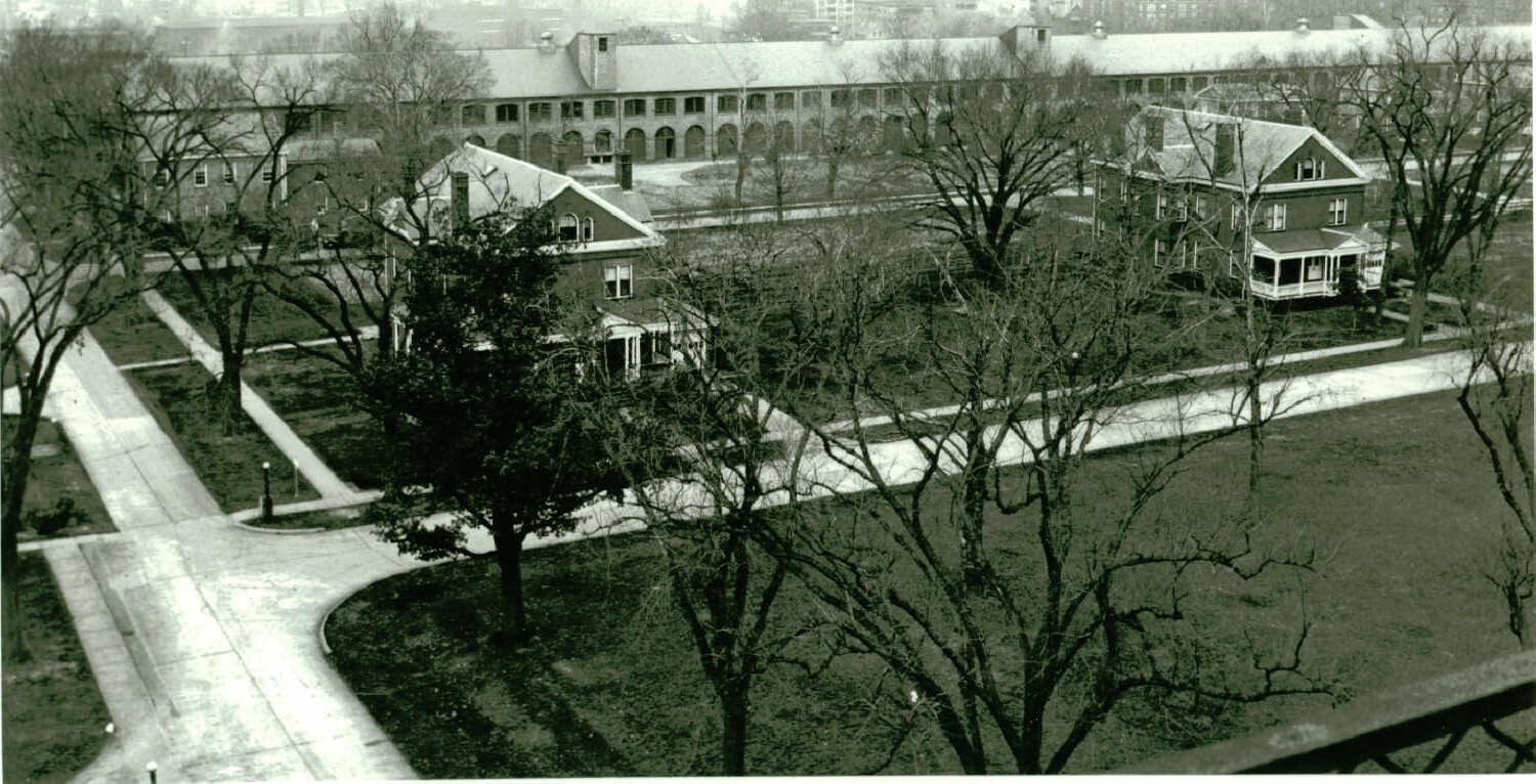 Black and white arial photo looking down on two houses with a large warehouse in the background. Paved streets and sidewalks run parallel to the houses and the warehouse. Trees partially obscure the house on the left from view.