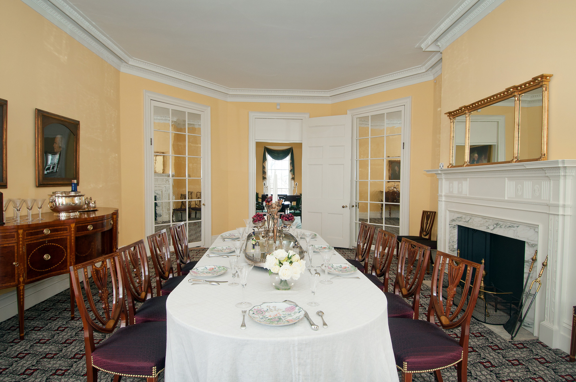 A table draped in a white cloth and set with china and silver ware, surrounded by mahogany chairs.