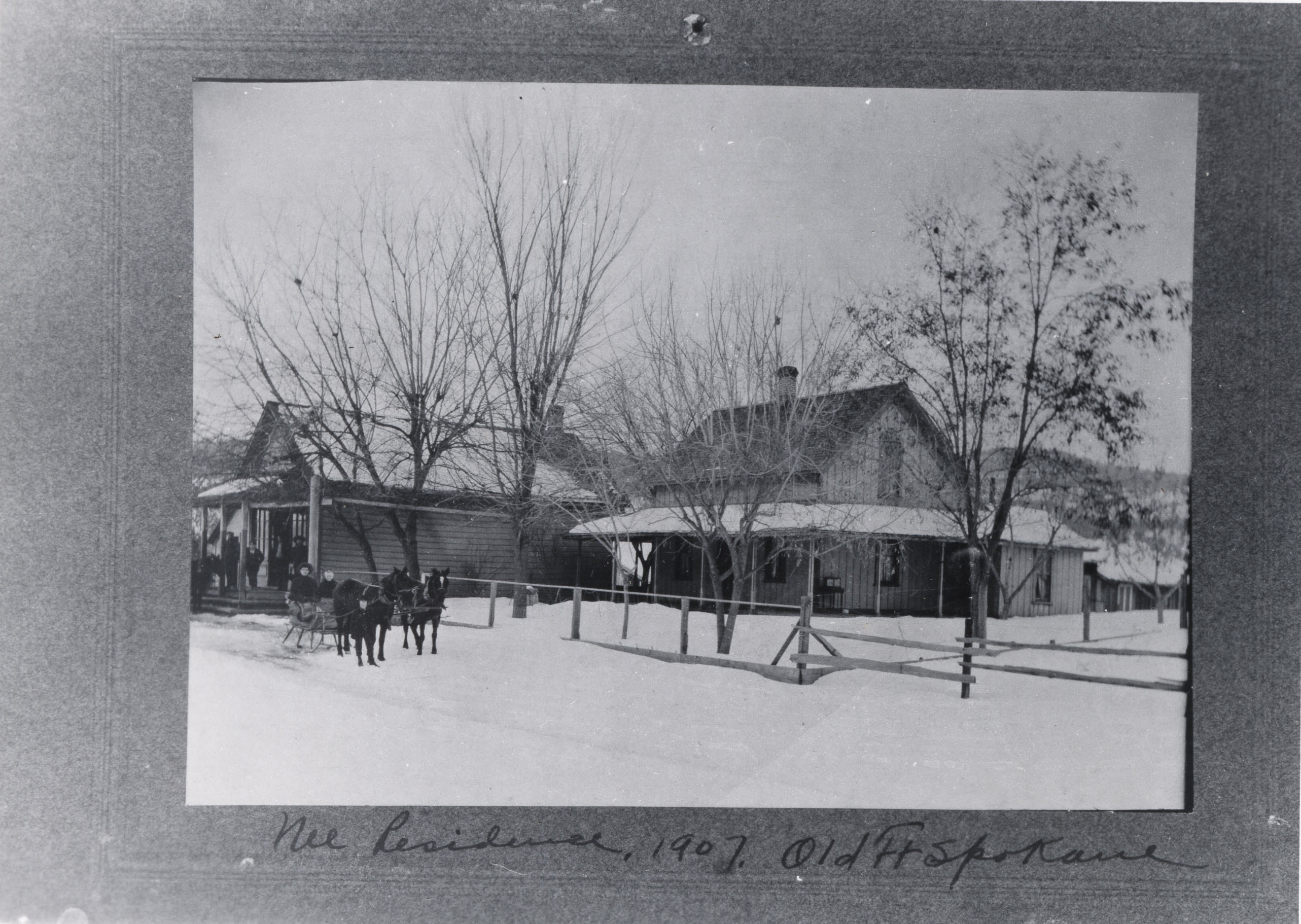 Black and white photograph of a complex of wooden buildings in the snow with horses pulling a sleigh