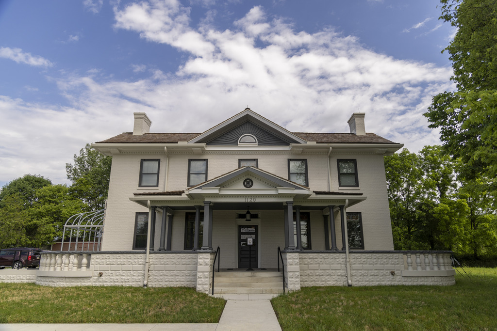 A large, two-story, light-colored home with five windows on the top level, peaked roofs on the first and second level in the center, lush green trees and grass around and behind the house, all under a partly cloudy sky.