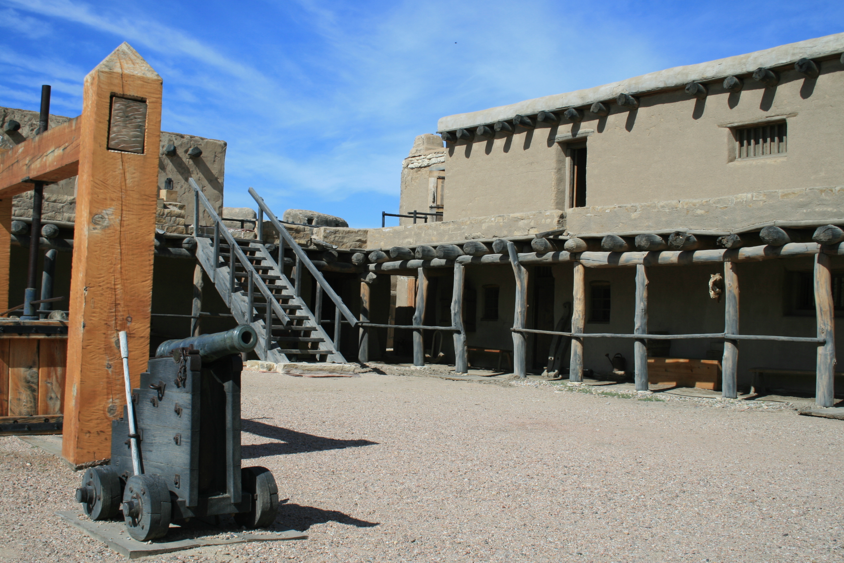 A historic adobe building with wooden beams, a staircase, and a cannon in the courtyard under a blue sky.