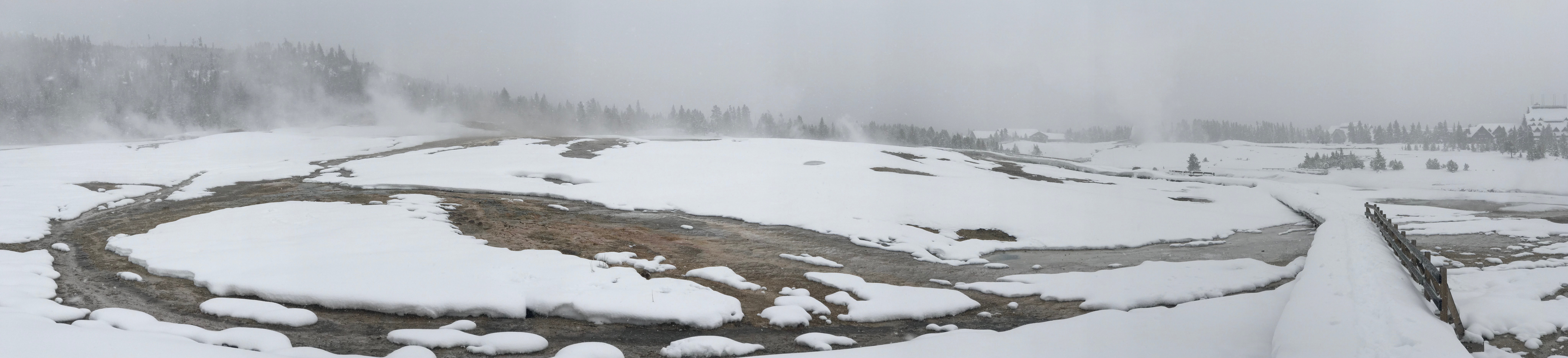Snow covers the hillside, with steam rising up from behind the hill and brown and orange water channels cut across the snowy scene.