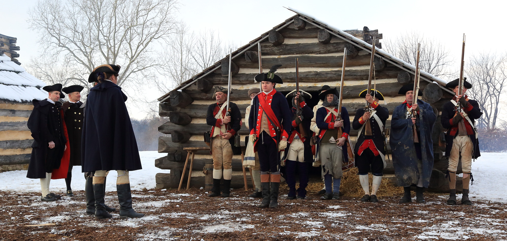 Twelve men wearing 18th century soldier uniforms stand in front of a log hut. Nine soldiers stand at attention holding muskets and three others stand in front, looking on.