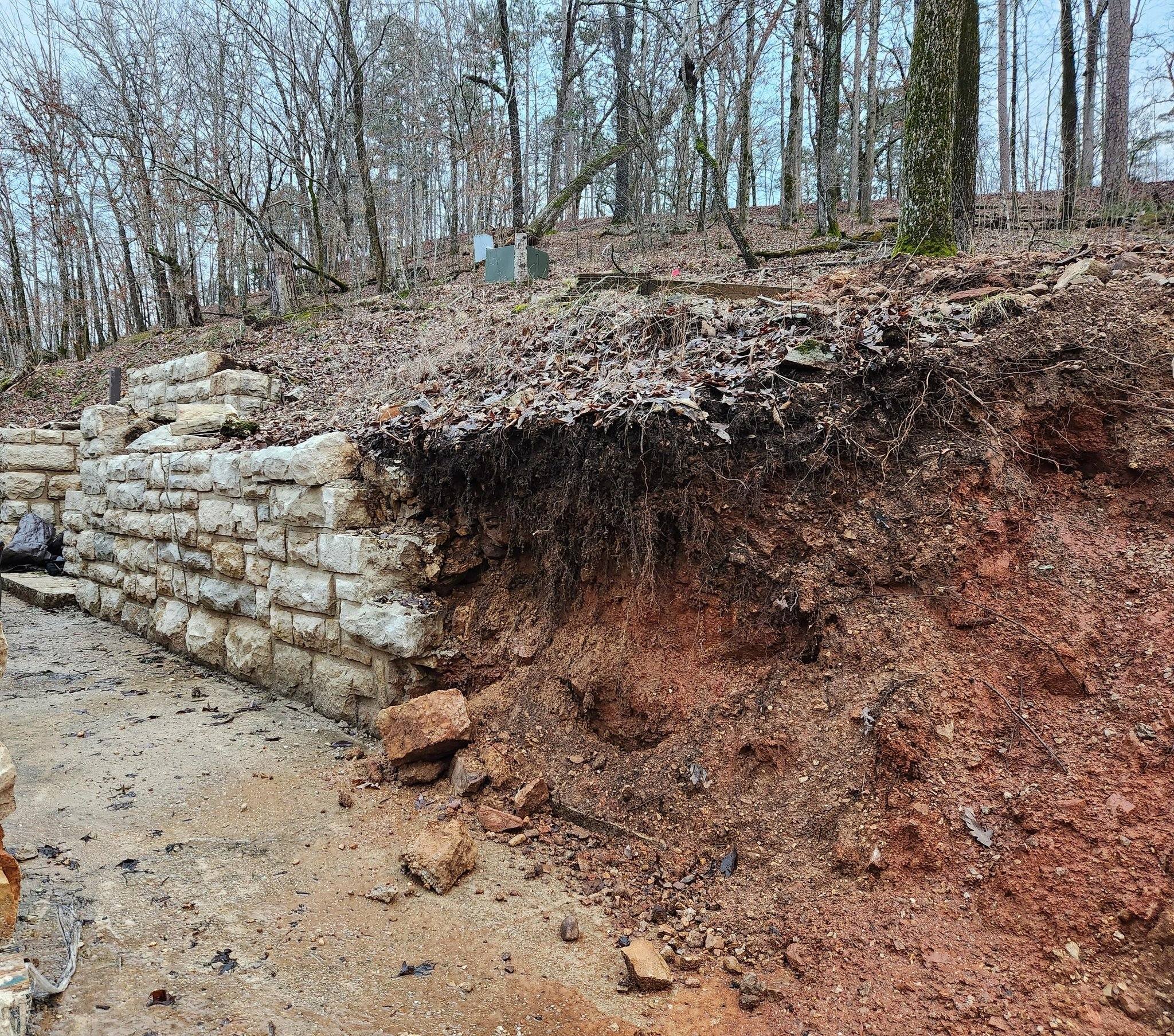 Part of the wall described in the second photo shot from farther down, with the inset containing the fountain just visible at the left of the frame. The wall extends to the center of the frame and then ends where the stones have been removed, revealing the red clay hillside with visible roots extending down from the top. Leafless trees with no undergrowth between them, indicating winter, can be seen on the hillside above, and blue sky is visible through the trees.