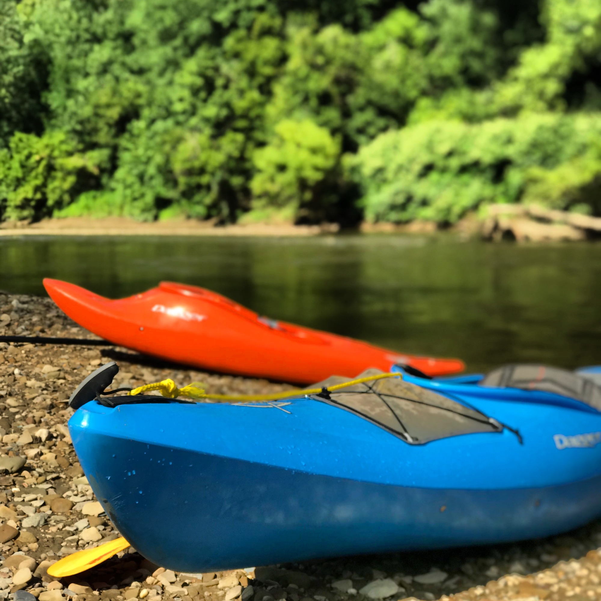 A blue and orange Kayak are in the foreground up on a bank next to the Cuyahoga River in summer.