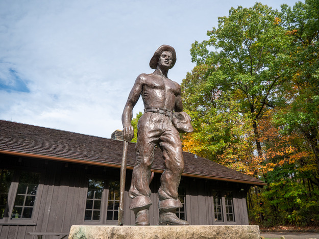 A bronze statue of a shirtless man wearing a wide-brimmed hat and leaning on the handle of on axe, outside a brown building.